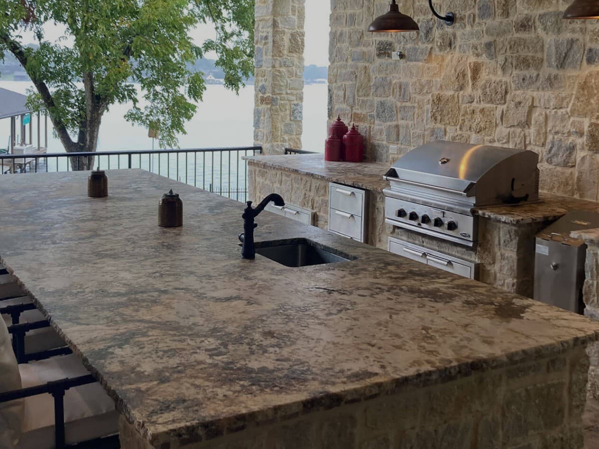 Finished outdoor kitchen with natural stone counters, sink, grill, and lake view